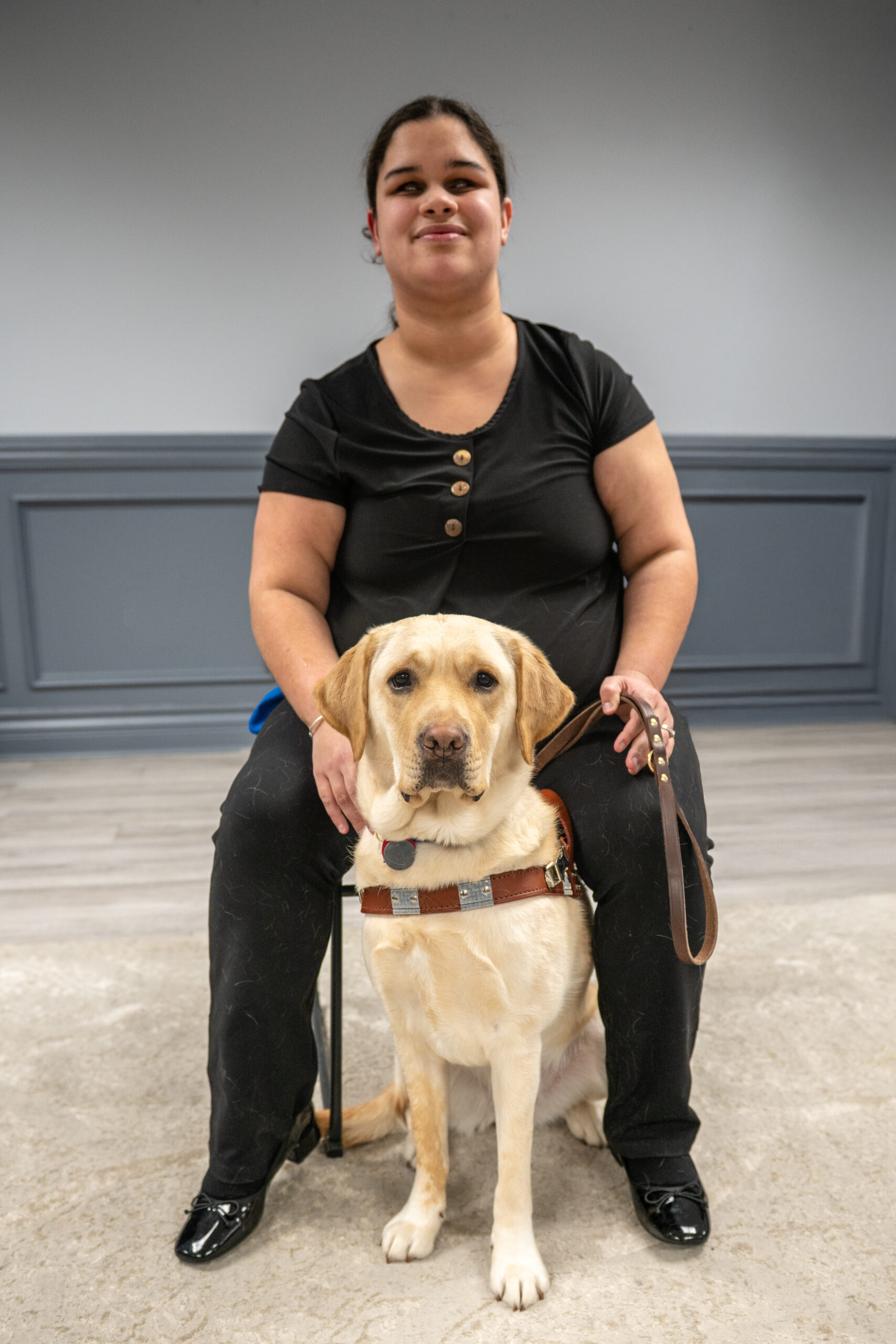 Chloe Walton sitting with her guide dog Odette, a yellow Labrador, in front of a grey wall. Chloe is wearing black and smiling at the camera.